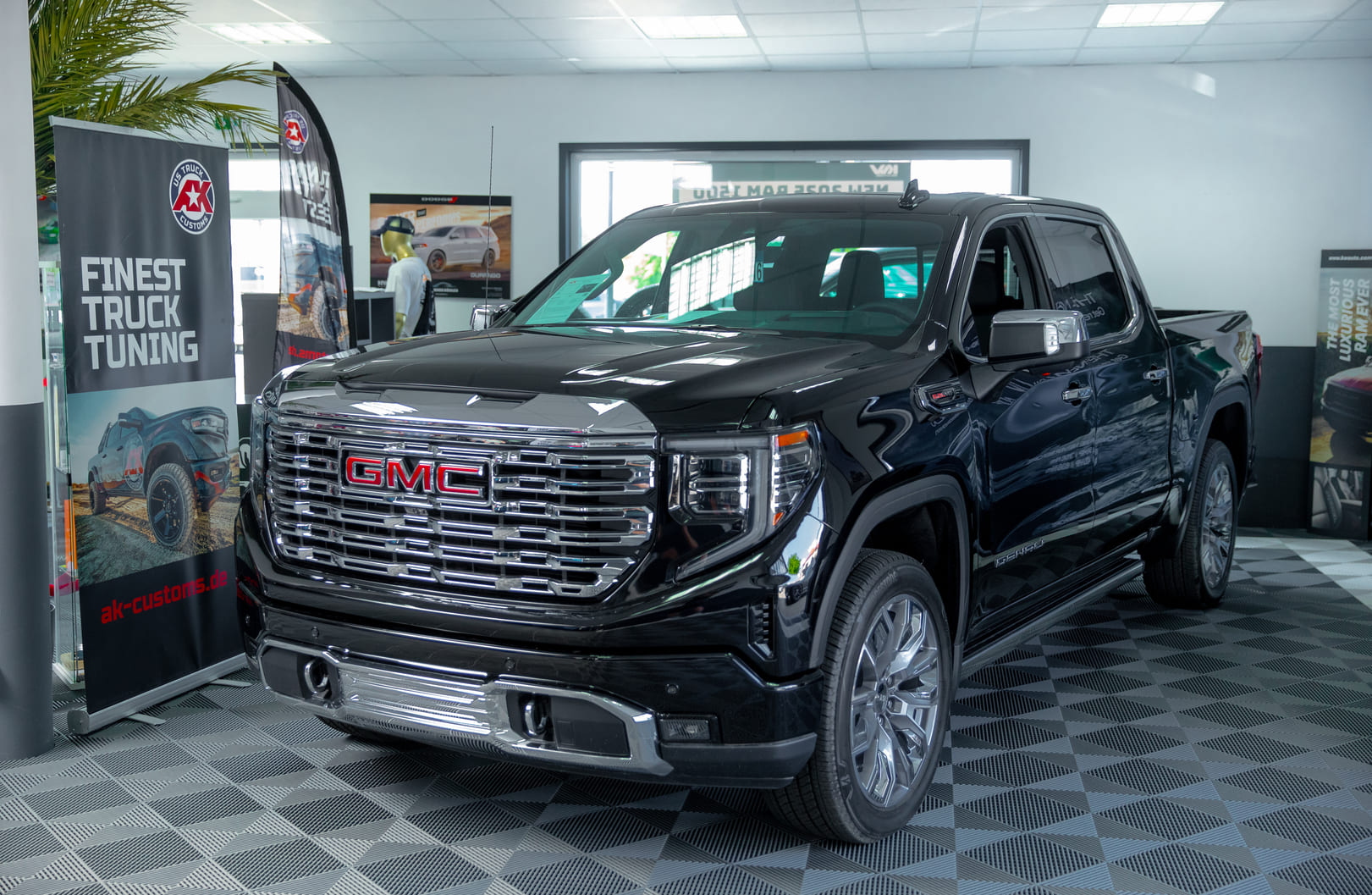Das Bild zeigt einen GMC Sierra 1500 Denali (Modelljahr 2025) in tiefschwarzer Lackierung, ausgestellt im Showroom von Auto Köhler. Der Truck beeindruckt mit seinem markanten, verchromten Denali-Kühlergrill, glänzenden 22-Zoll-Felgen und edlen Chromakzenten an Front, Spiegeln und Stoßfängern. Im Hintergrund sind Banner von AK Customs zu sehen, die auf individuelles Truck-Tuning hinweisen – passend zum hochwertigen Premium-Ambiente der Ausstellungsfläche.